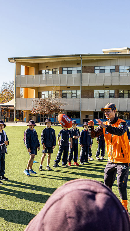 Mother Teresa Primary Westmead students on sports field. The students are playing AFL with a coach.