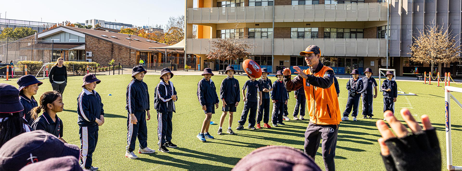 Mother Teresa Primary Westmead students on sports field. The students are playing AFL with a coach.
